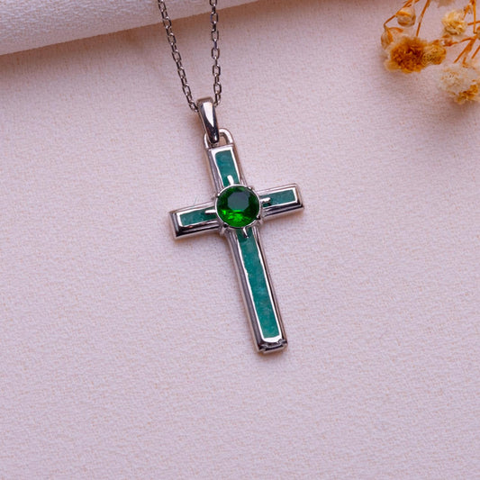 Silver Birthstone Cross Necklace with a green round center stone and green rectangular accents on pale fabric