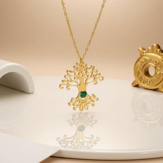 Gold Plated Silver Tree of Life pendant with curled branches, scrolling roots, and a green center stone on a cream display surface