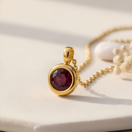 Gold Plated Silver Birthstone Pendant Necklace with a round deep red stone on a pale textured surface