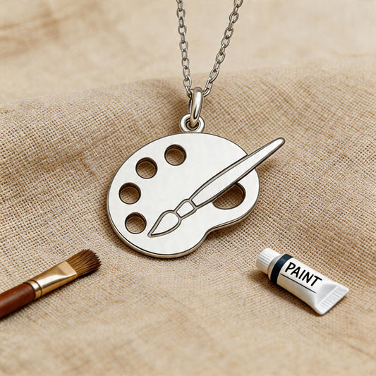 Close view of the Painter Palette Necklace on textured beige fabric beside an artist paintbrush and paint tube